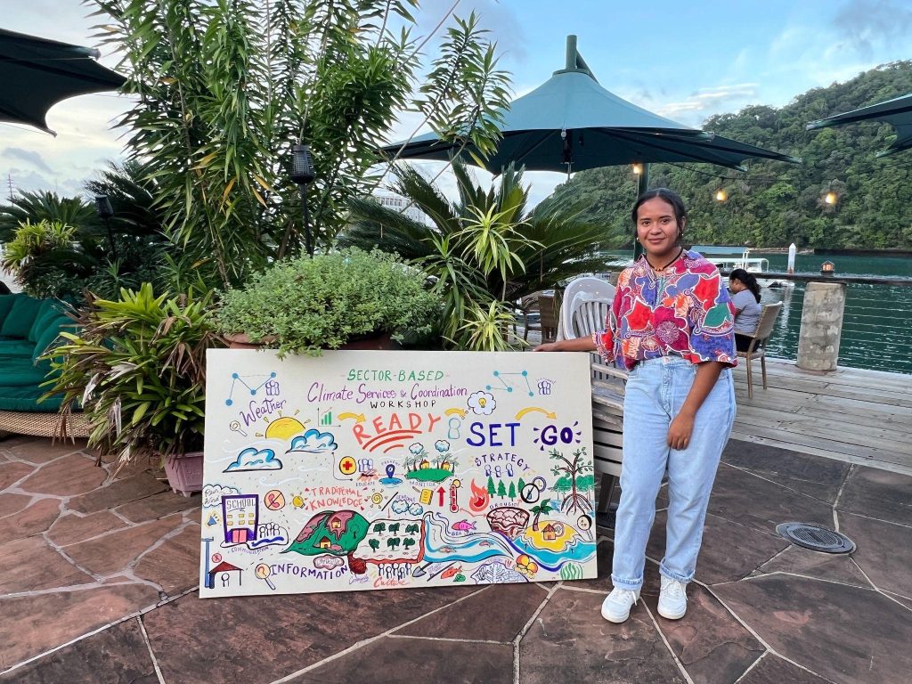 woman poses next to a hand-drawn sign for the workshop in Palau