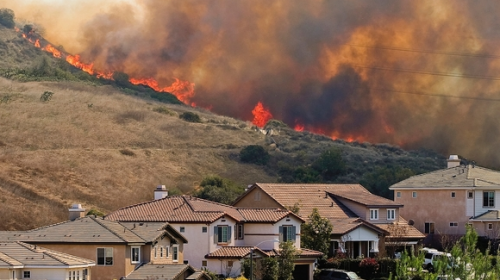 Houses in the foreground at the bottom of a hill with fire and smoke coming over the ridge