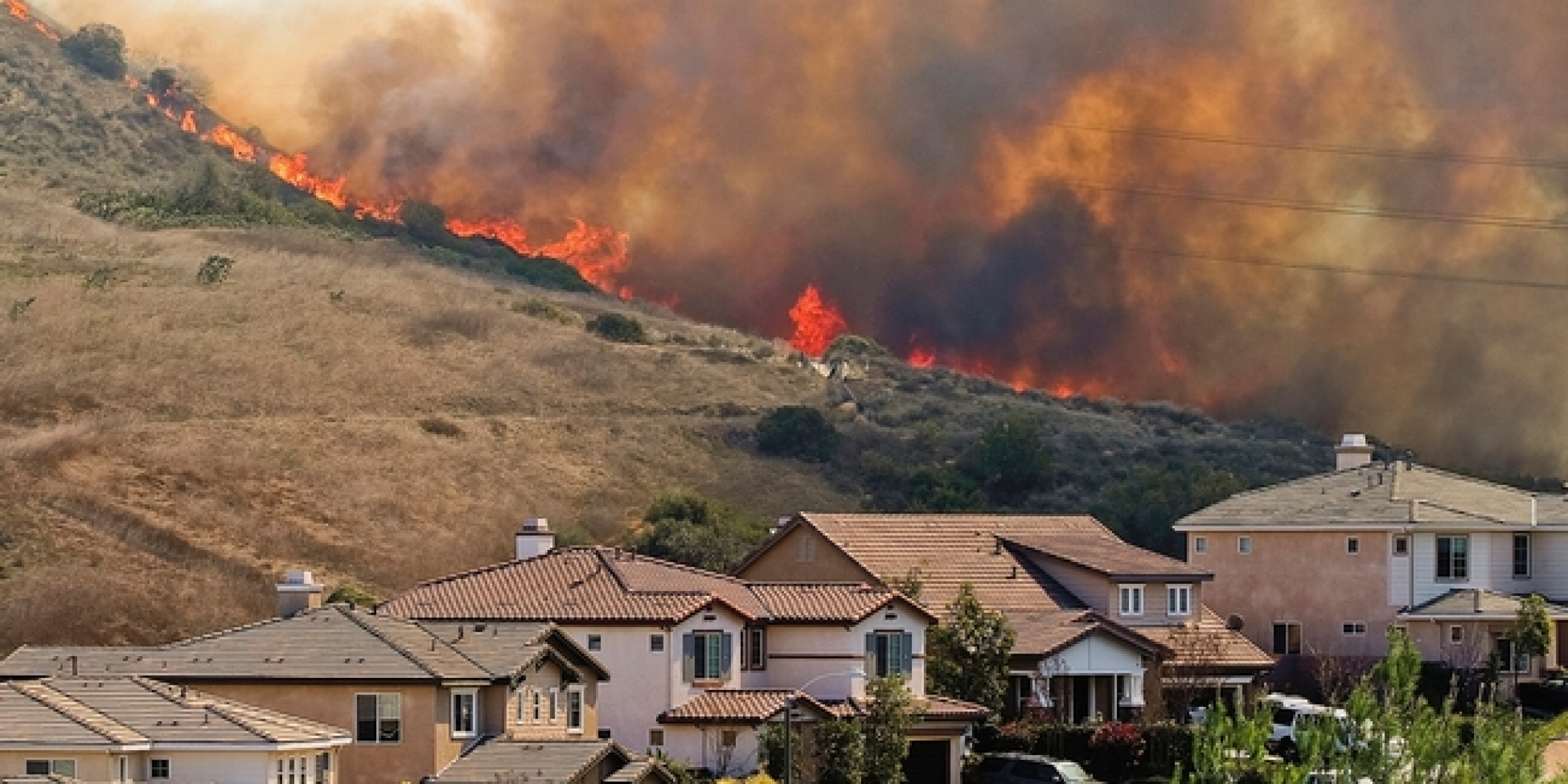 Houses in the foreground at the bottom of a hill with fire and smoke coming over the ridge